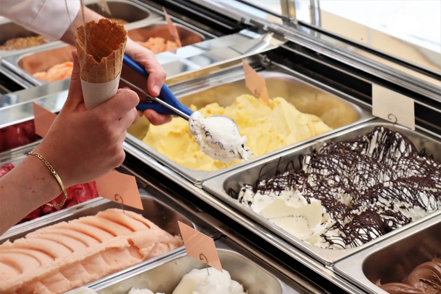 Person holding an empty waffle cone while scooping white and chocolate drizzled ice cream from a display case with various ice cream flavors visible.