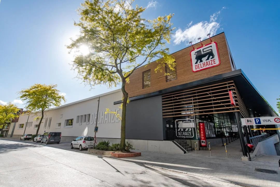 Modern Delhaize supermarket building with wooden and white facade, parked cars, and trees under a clear blue sky with sun.
