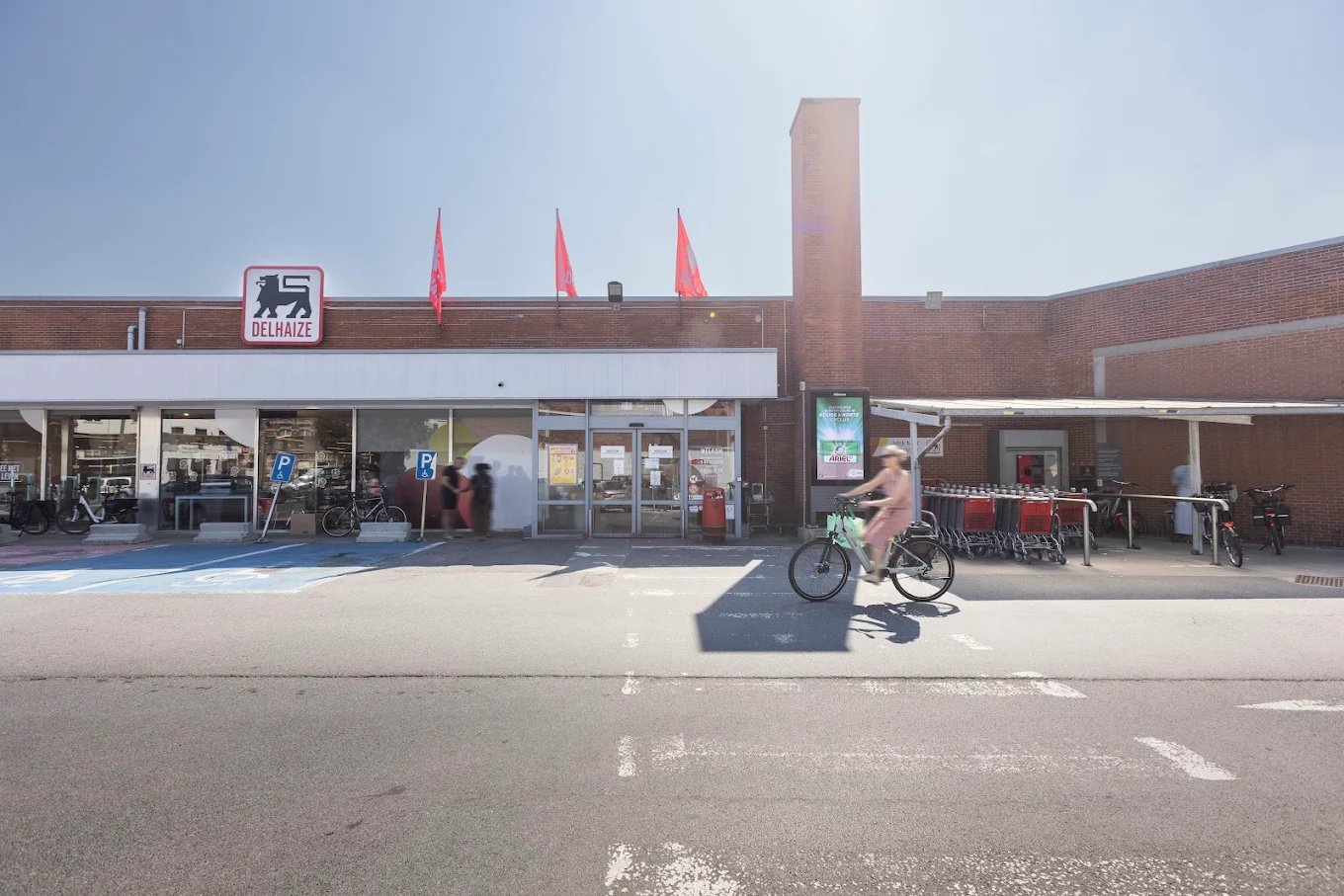 Front entrance of a Delhaize supermarket with red flags on the roof and a person riding a bicycle in front.
