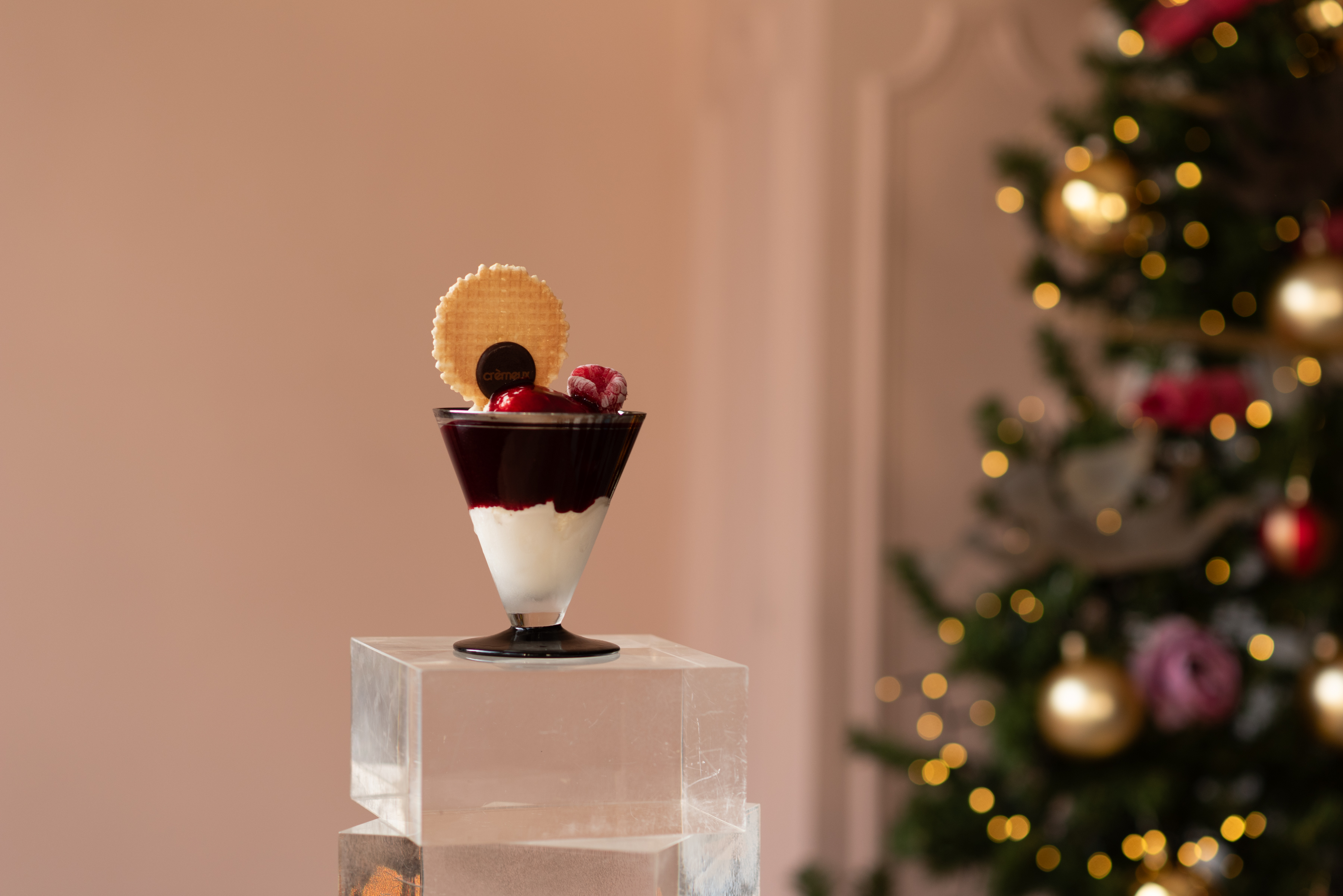 Layered dessert in a conical glass topped with a wafer, raspberry, and chocolate disc, placed on a clear acrylic pedestal with a decorated Christmas tree in the background.