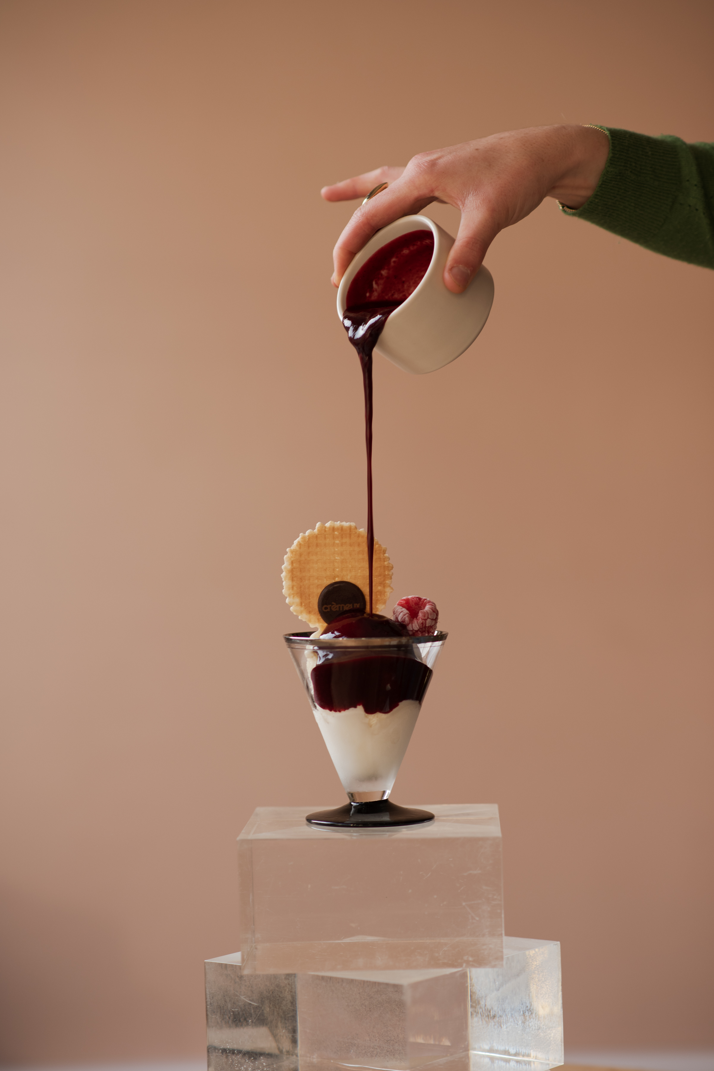 Hand pouring dark red sauce from a white cup onto vanilla ice cream topped with a wafer and raspberry in a glass dessert cup.