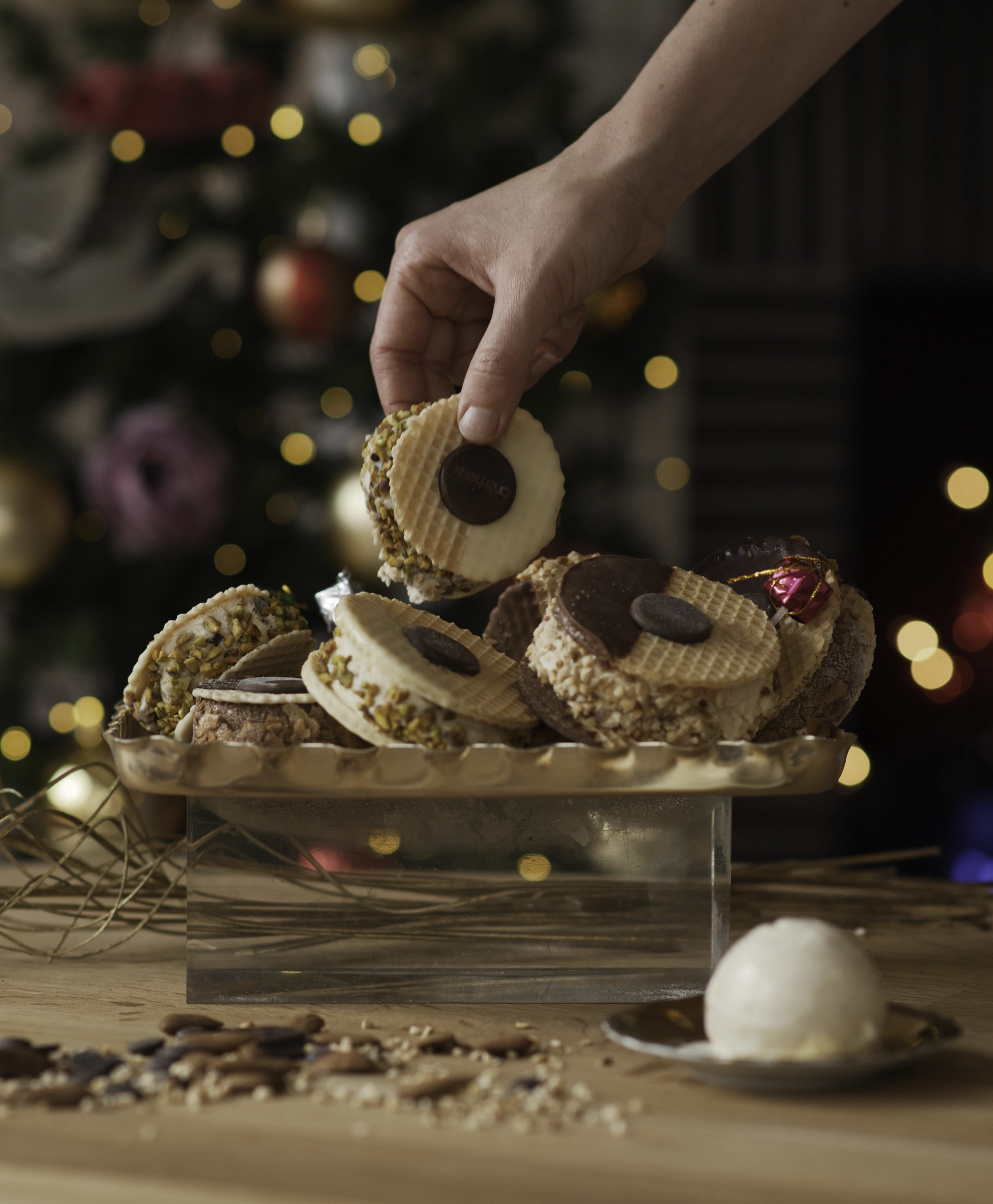 Hand picking a wafer cookie from a tray filled with various decorated wafer cookies, with a blurred Christmas tree in the background.
