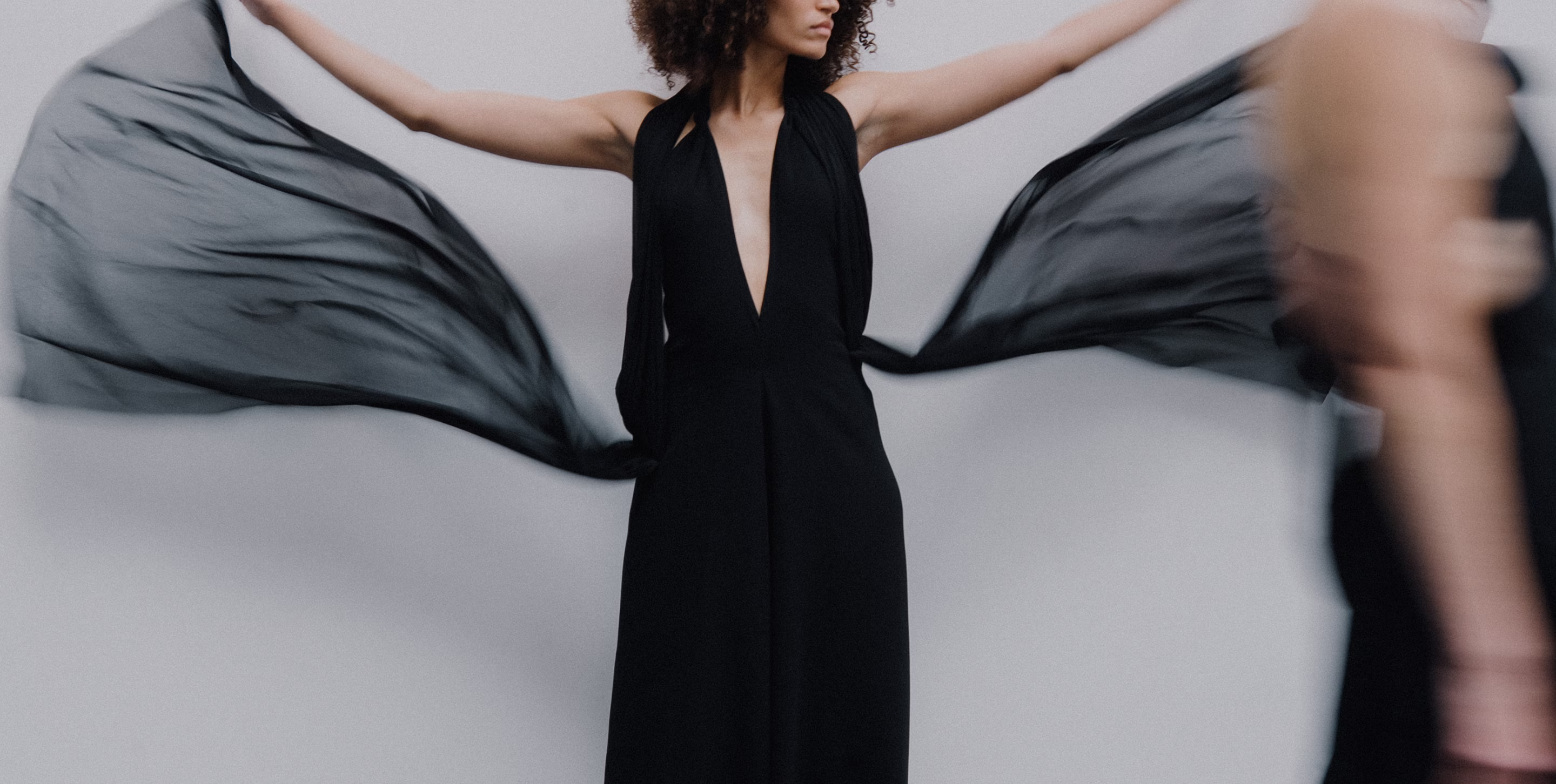 Woman in a black dress spreading sheer black fabric with arms outstretched against a plain background, with a blurred figure passing by.