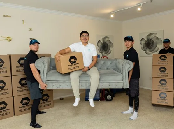 Team of professional movers lifting a sofa while holding branded moving boxes inside a clean, furnished living room, showcasing Payable Moving’s residential moving services.