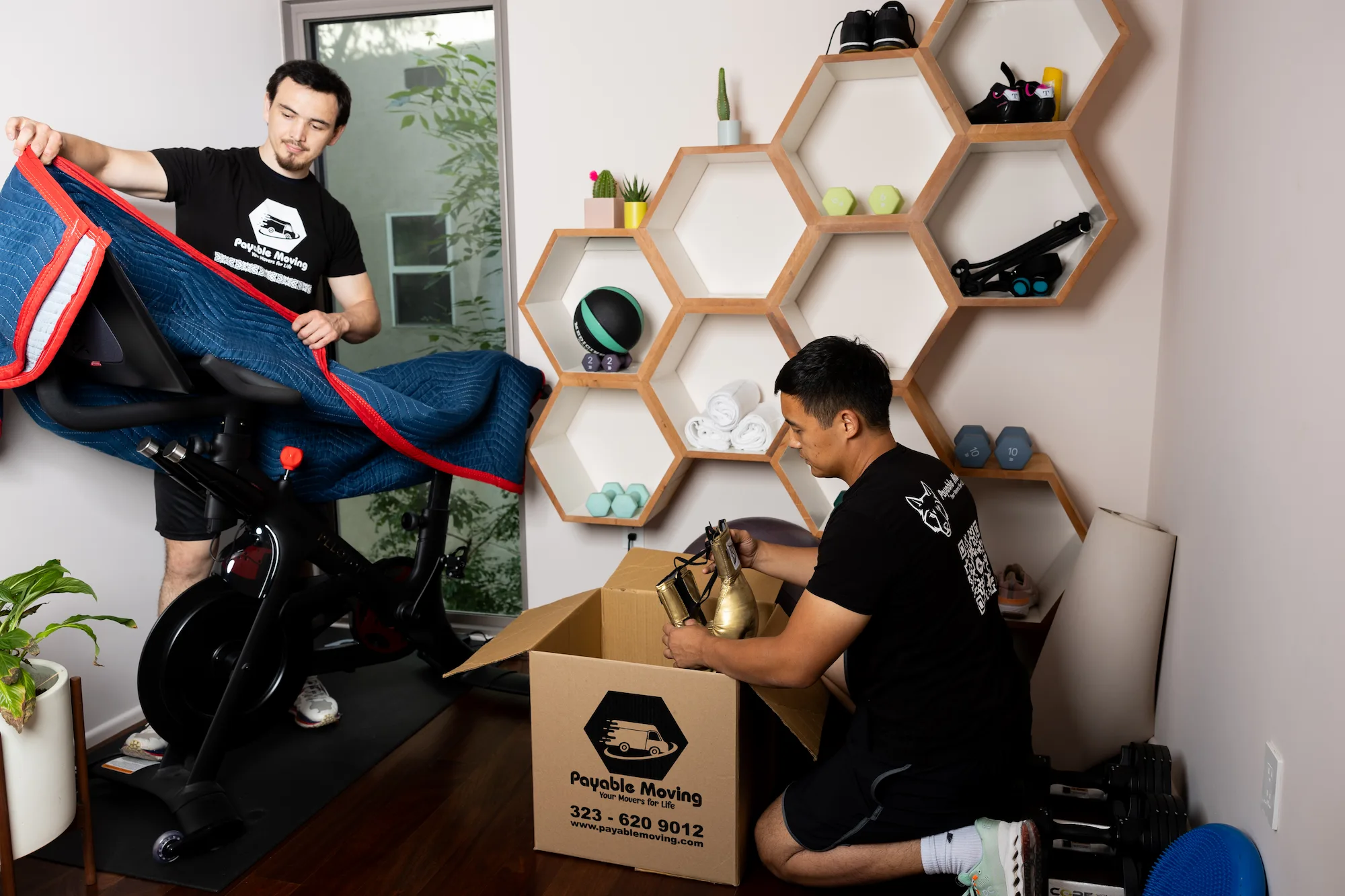 Professional movers packing fitness equipment in a gym space, with one worker wrapping a stationary bike and another boxing dumbbells, showcasing Payable Moving’s commercial moving services.