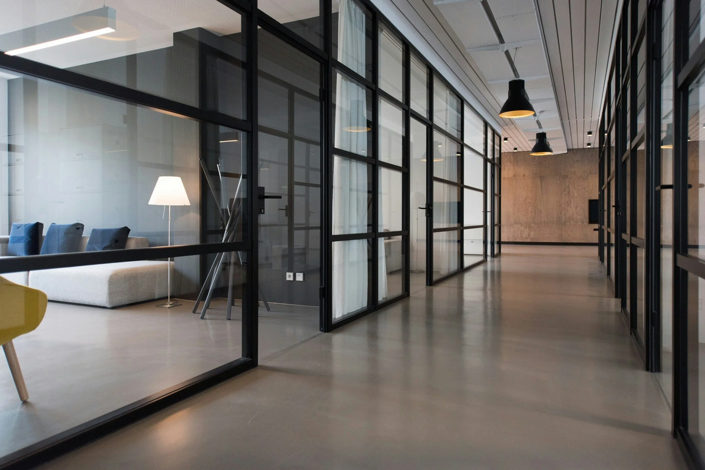 Modern office hallway with glass-walled meeting rooms, black metal frames, polished concrete floors, and warm pendant lighting, with a lounge area visible on the left.
