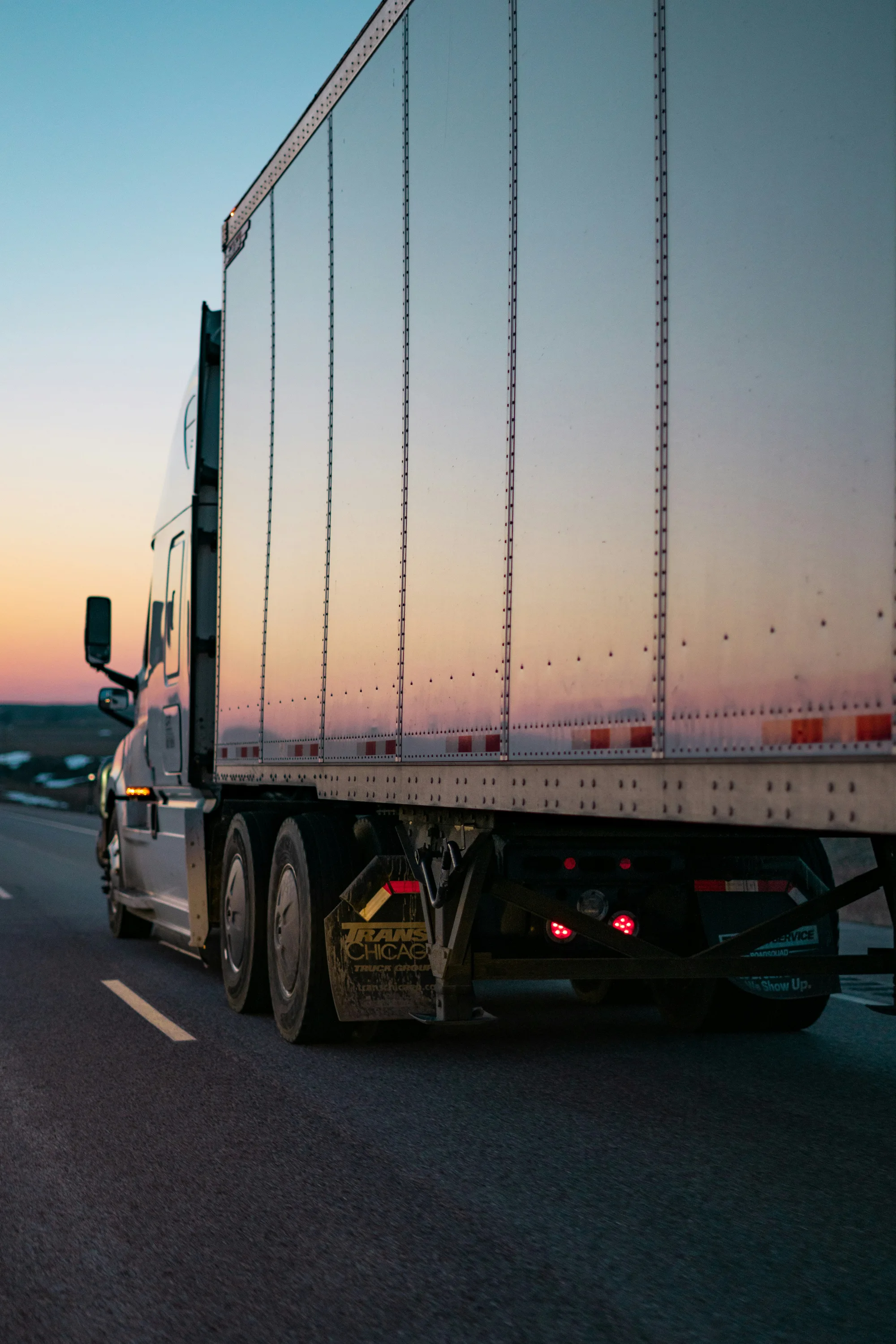Close-up side view of a large semi-truck driving down a highway at sunset, with warm sky colors reflecting on the trailer.