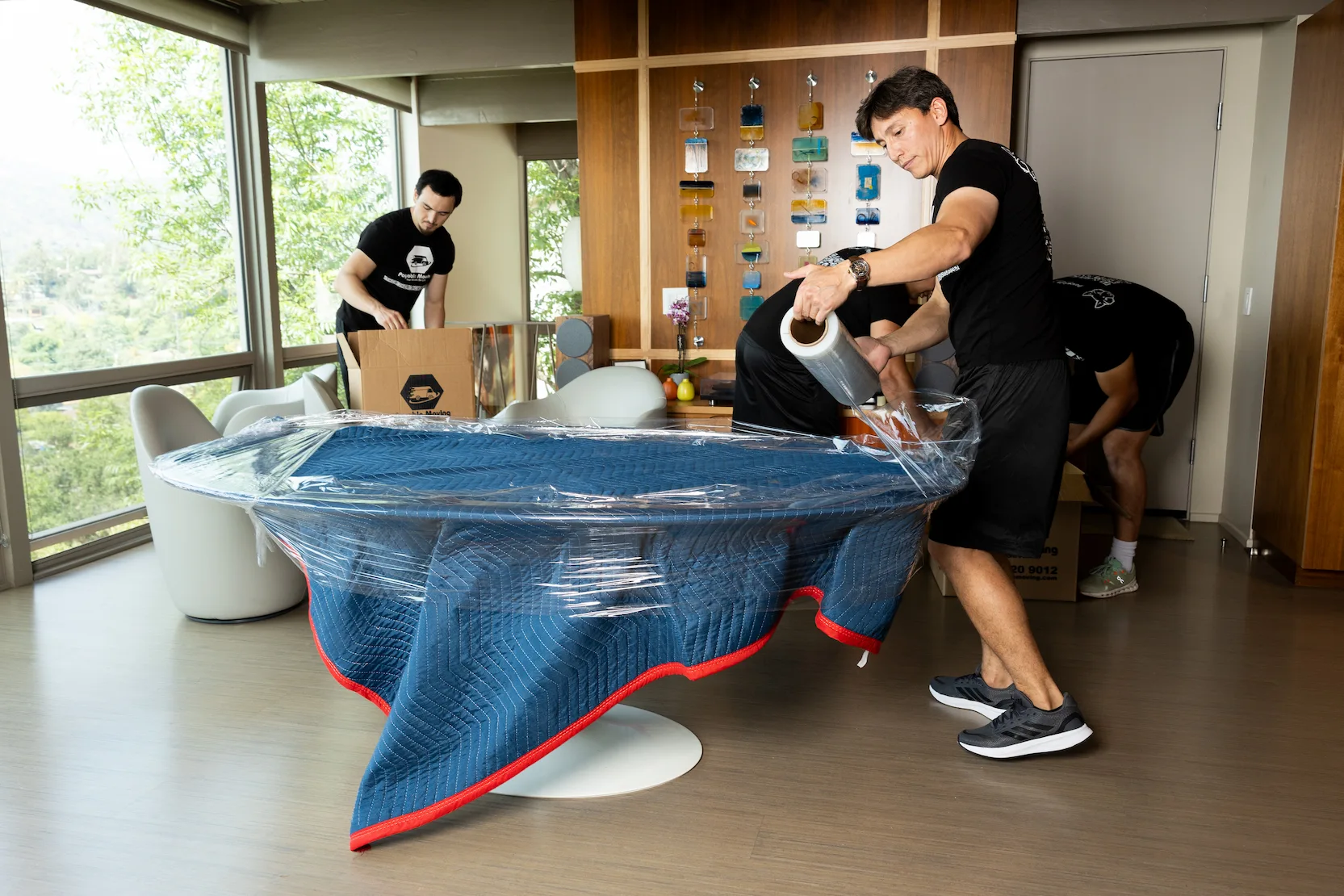 Payable Moving team professionally wrapping a large dining table with protective blankets and plastic wrap while preparing items for relocation inside a modern home.