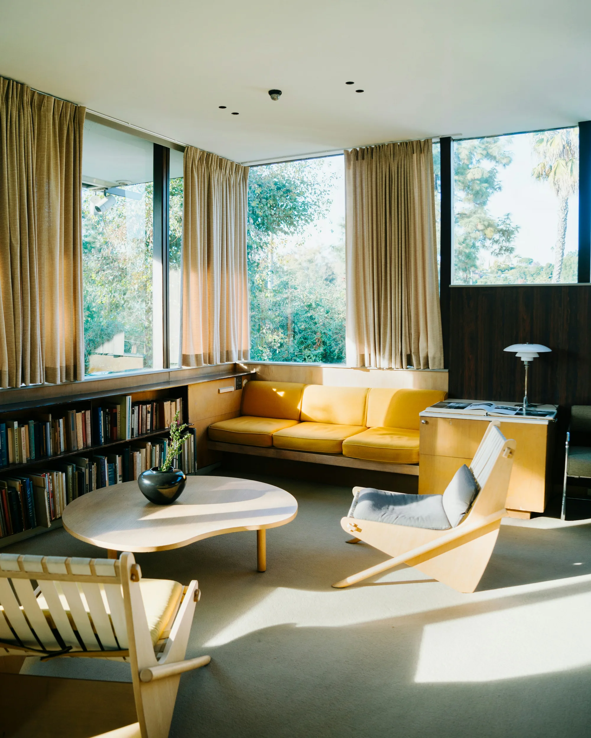 Bright mid-century modern living room with floor-to-ceiling windows, yellow sofa, wooden furniture, and built-in bookshelves