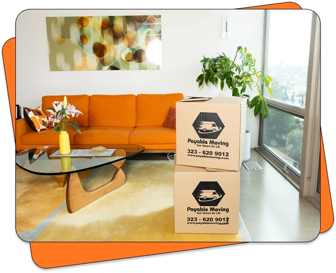 Living room with an orange sofa, glass coffee table, and two stacked Payable Moving cardboard boxes placed near a large window with natural light.