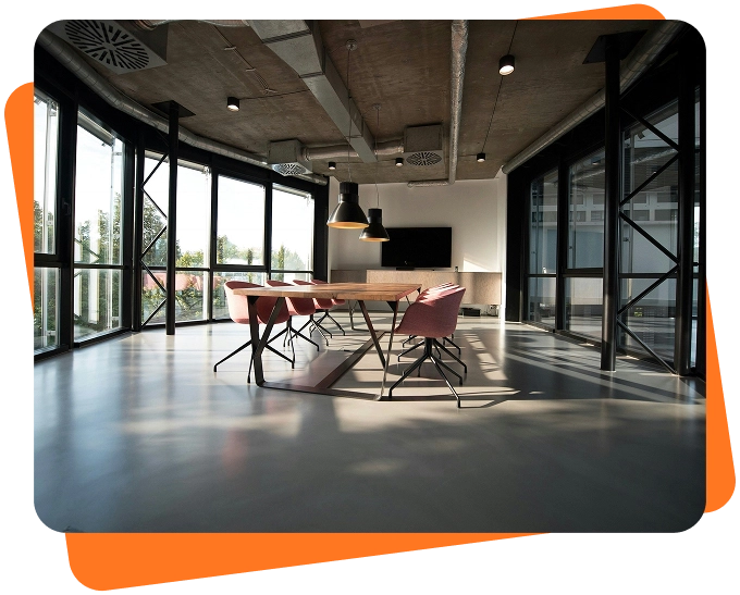 Modern conference room with large floor-to-ceiling windows, a wooden meeting table, pink chairs, and industrial ceiling fixtures letting in natural daylight.