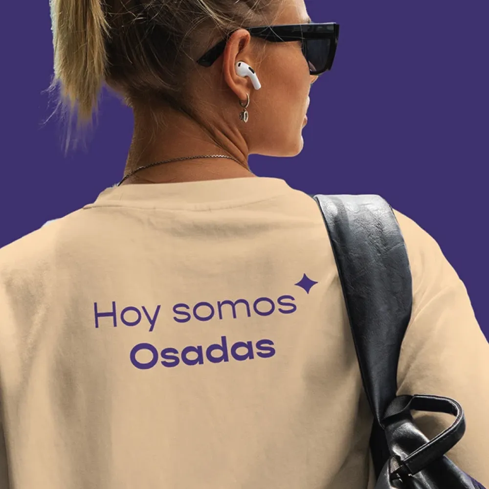 Mujer de espaldas con gafas oscuras, auriculares blancos, mochila negra y camiseta beige con la frase “Hoy somos Osadas” en letras moradas, manteniendo coherencia sistema visual de la marca. Servicio de branding, ubicación Perú.