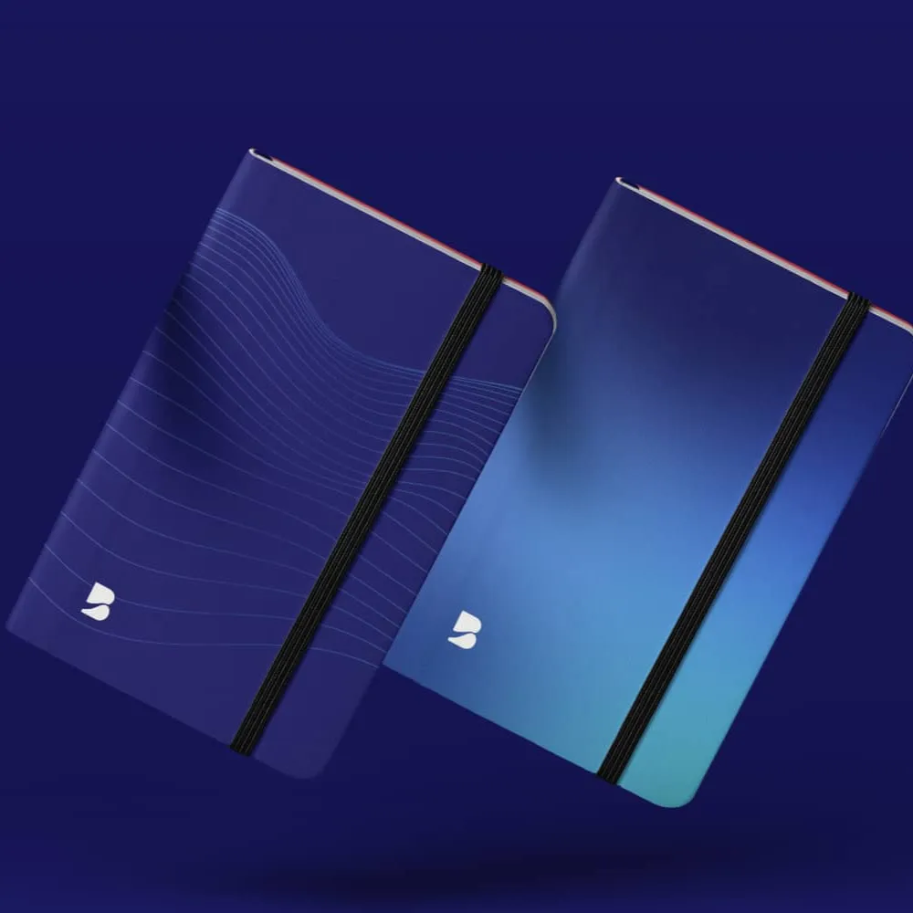 Dos cuadernos azules con bandas elásticas negras; uno con diseño de líneas onduladas y otro con degradado de azul claro a oscuro, manteniendo coherencia con el sistema visual de la marca. Diseño de identidad, ubicación Madrid, España.