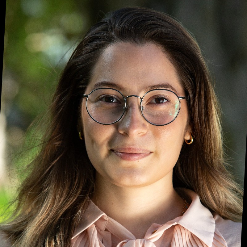 Mujer joven con cabello largo y suelto, usando lentes redondos y una blusa rosa clara, sonriendo ligeramente frente a un fondo desenfocado natural.