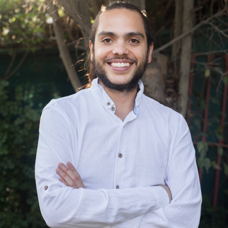 Hombre con cabello recogido y barba, sonriendo con brazos cruzados, usando camisa blanca en un entorno al aire libre.
