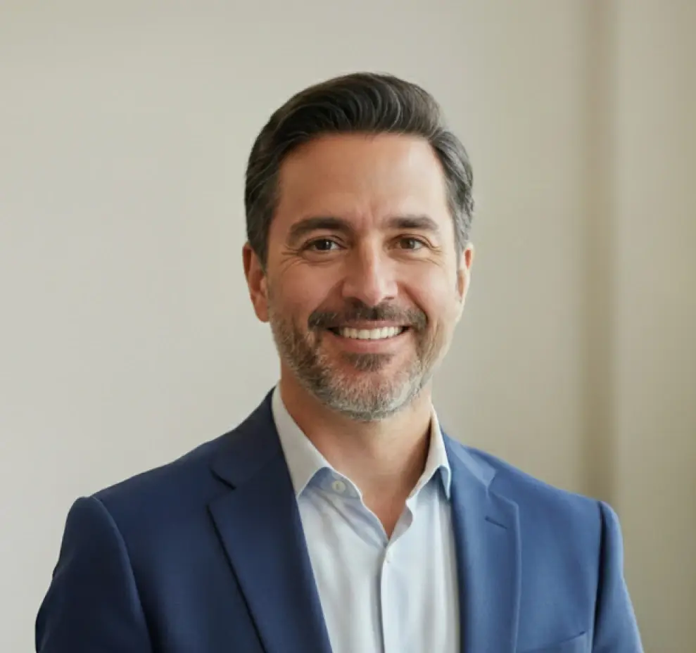 Hombre sonriente de cabello oscuro y barba canosa vestido con saco azul y camisa blanca.