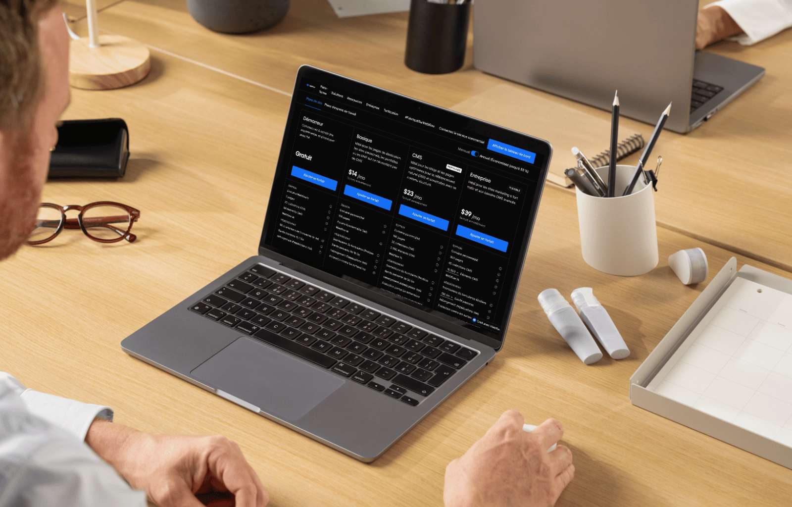 Un homme regarde un ordinateur portable affichant une page de tarification de services avec plusieurs options d'abonnement sur un bureau en bois clair.