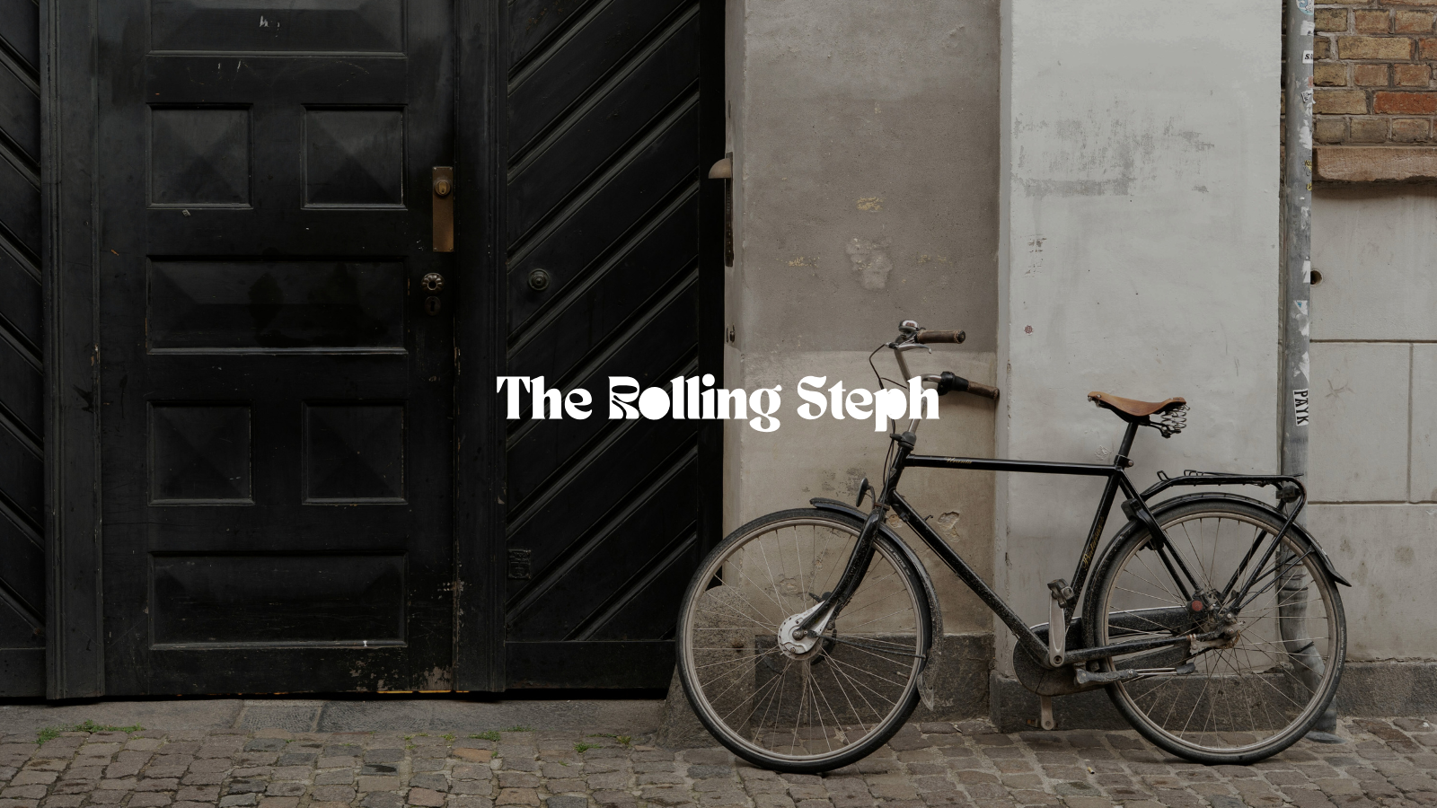 Vélo noir appuyé contre un mur beige à côté d'une porte en bois noire sur un trottoir pavé.