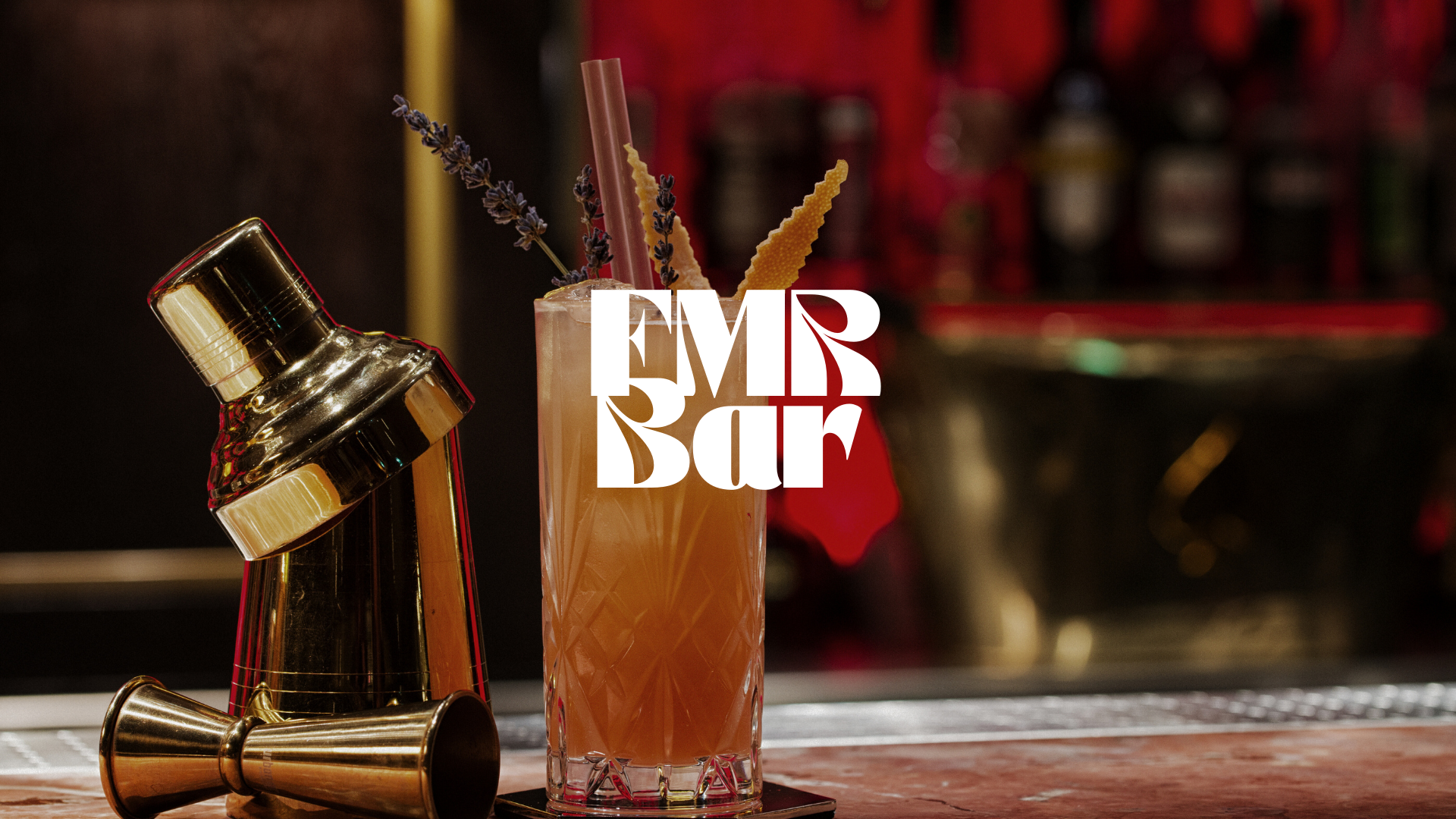 Cocktail décoré de brins de lavande et de zestes d'agrume à côté d'un shaker et d'un jigger en métal doré sur un bar.