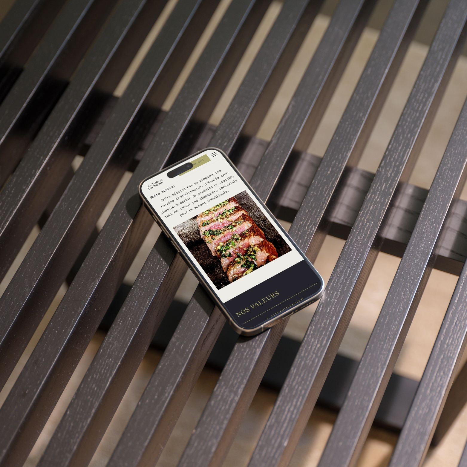 Un smartphone posé sur un banc en bois affiche une page de site Web avec une photo de steak grillé tranché et un texte en français sur la mission d'un restaurant.