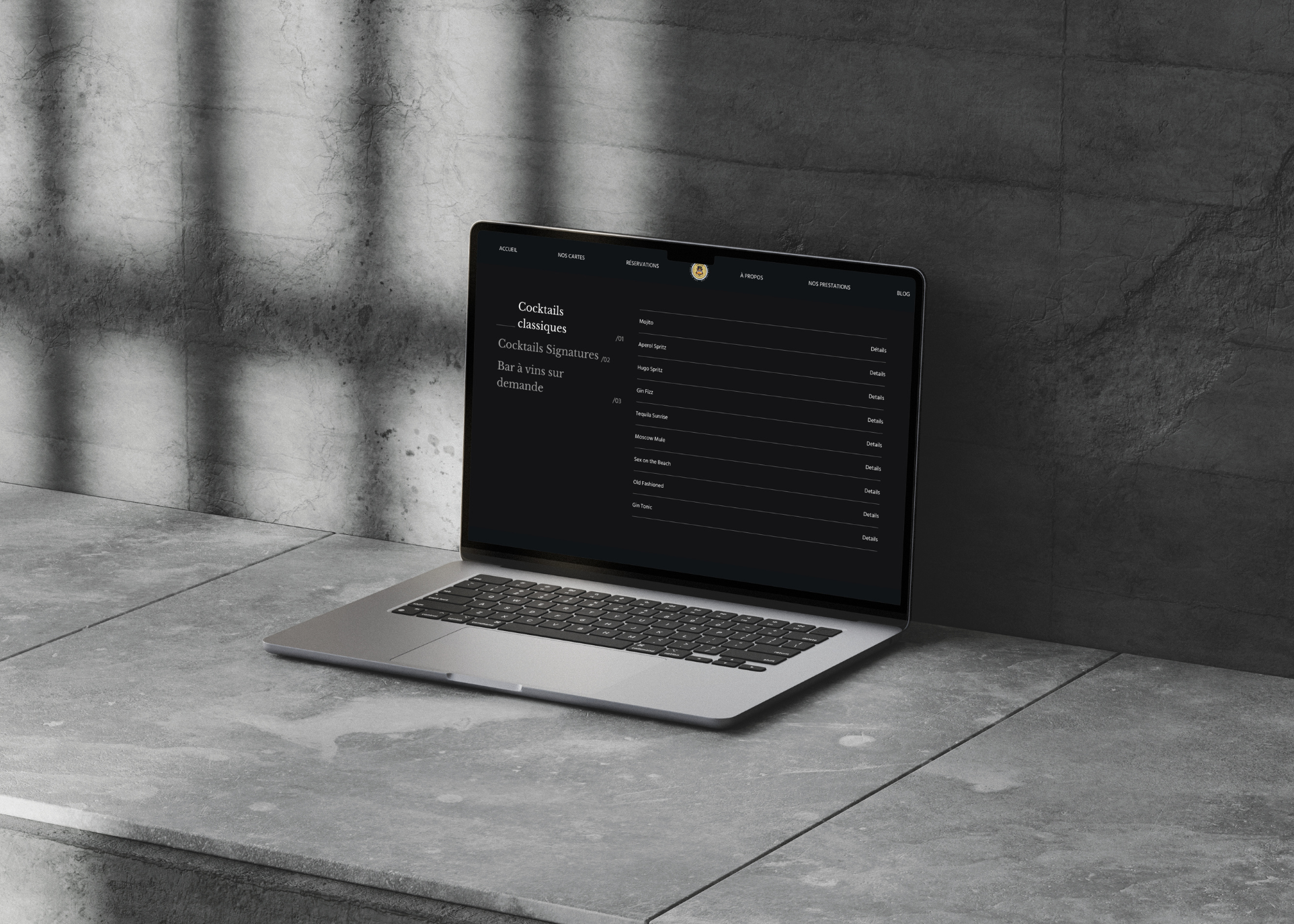 Ordinateur portable ouvert affichant un menu de cocktails classiques avec une liste de boissons sur un fond sombre, posé sur un comptoir en béton gris sous une lumière tamisée.