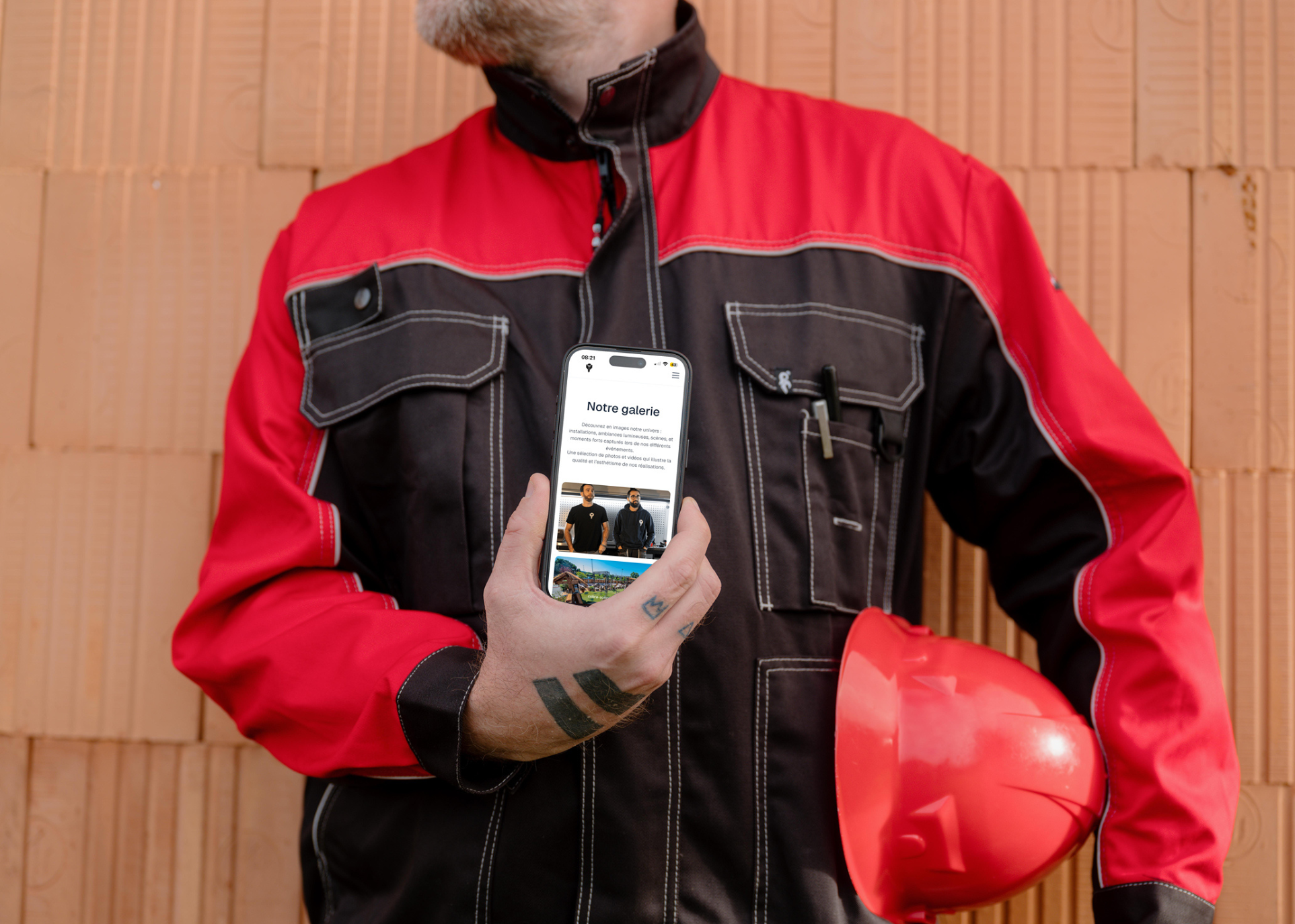 Ouvrier en veste rouge et noire tenant un casque rouge et un smartphone affichant une galerie de travaux en construction.