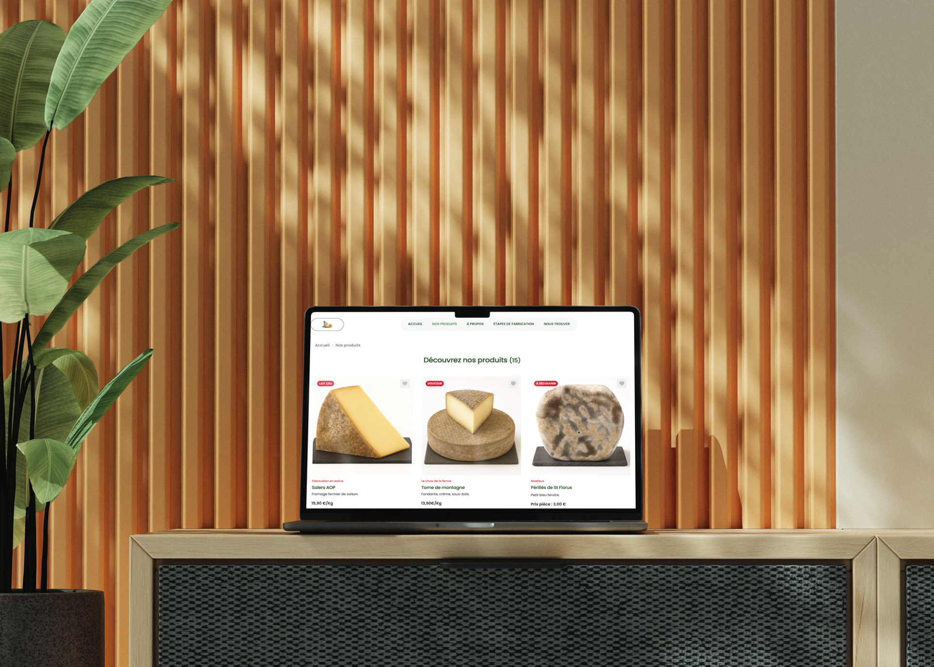 Ordinateur portable affichant une page web avec trois fromages différents sur un meuble devant un mur de lattes en bois, une plante verte à gauche.