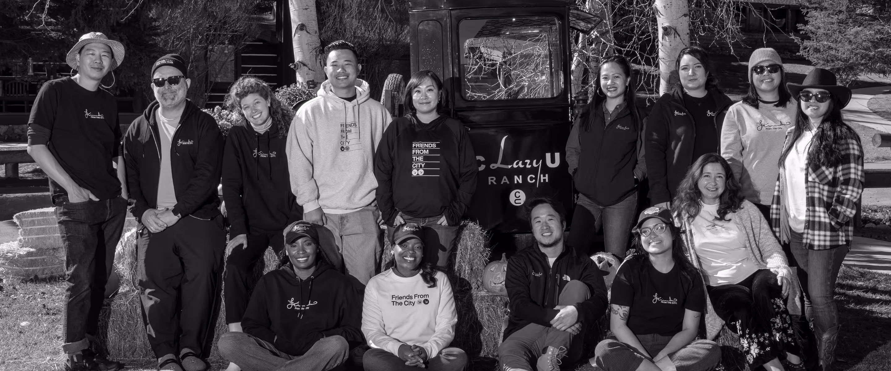 Group portrait of diverse people outdoors in front of a ranch sign, some sitting on hay bales and others standing, smiling and wearing casual clothes and hats.
