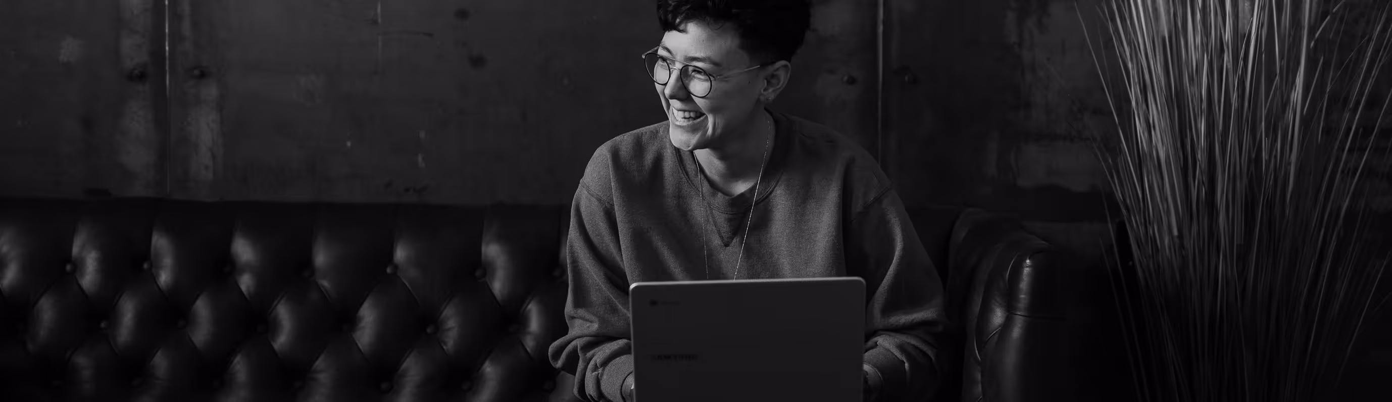 Person with glasses sitting on a tufted leather couch, smiling while using a laptop.
