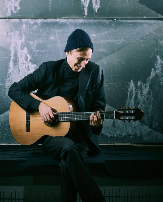Jakob Heymann sitzt mit einer Gitarre in seiner Hand und lächelt.