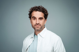 Oliver Wnouk lächelt in die Kamera vor einem hellblauen Hintergrund 