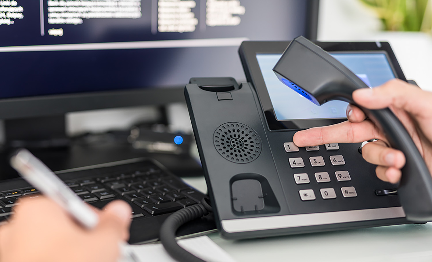 Hand holding a phone handset and pressing a button on a modern office desk phone with a touchscreen, keyboard and computer monitor in the background.