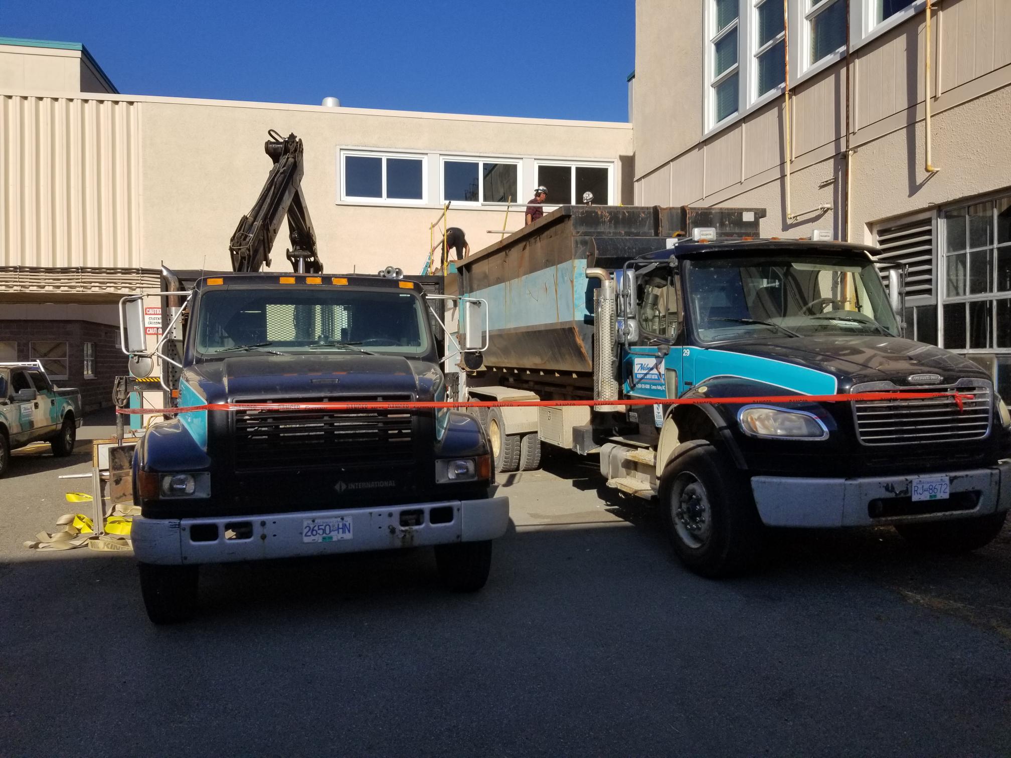 crane truck loading a metal debris bin onto a flatbed trailer at a job site
