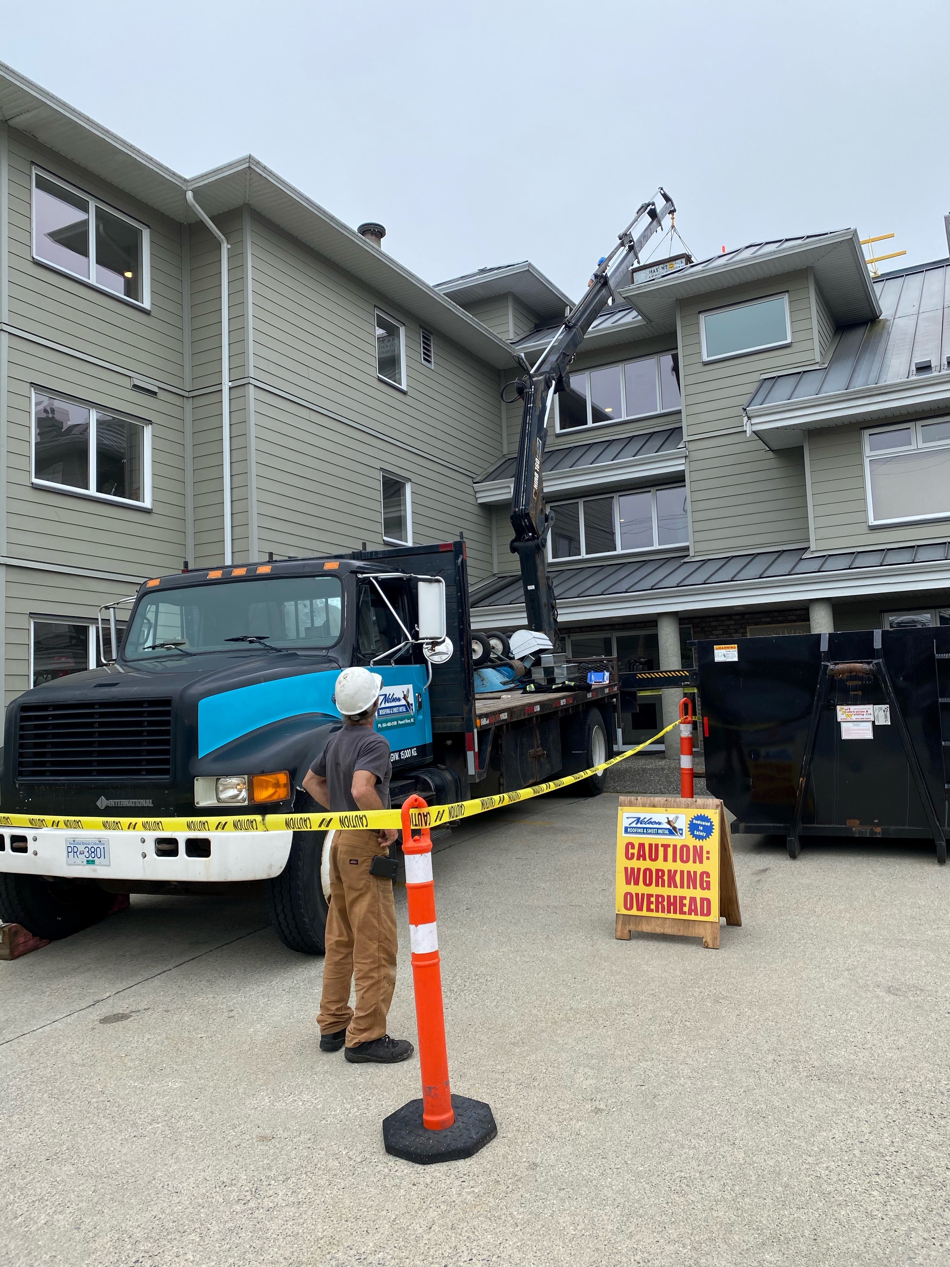 boom truck crane lifting roofing material onto a multi-story building, worker and caution signage below