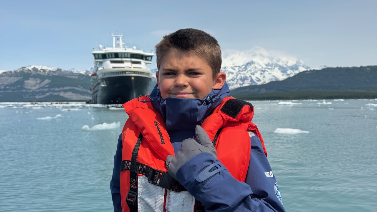 Colt Lockwood sits on a Zodiac boat in an Alaskan fjord with an expedition cruise ship in the background