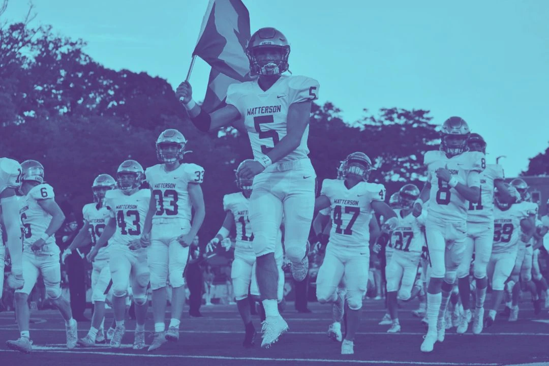 photo of an american football team entering the field, with the captain of the team carrying a flag