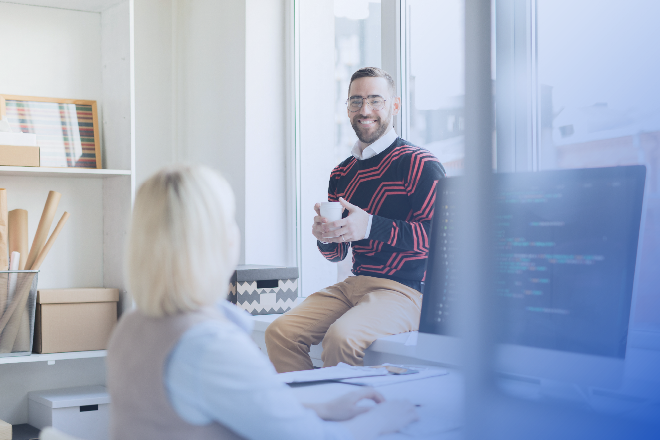 Man sitting on a windowsill, talking to a women working on a computer 