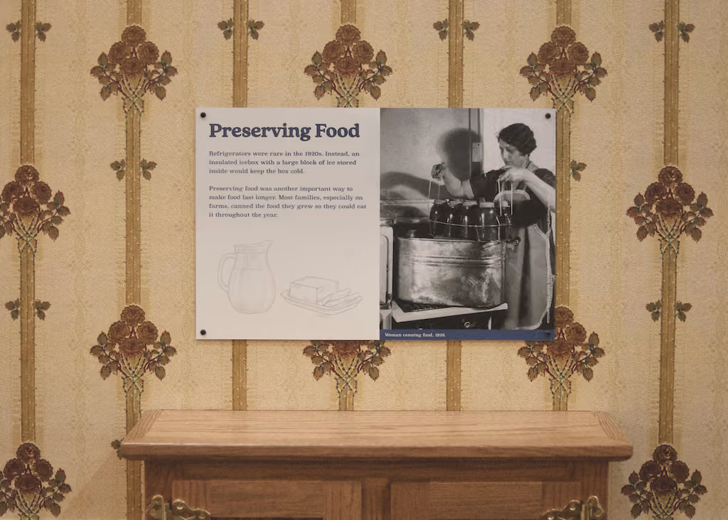 Display board titled 'Preserving Food' with 1920s canning photo on vintage wallpaper above wooden dresser.