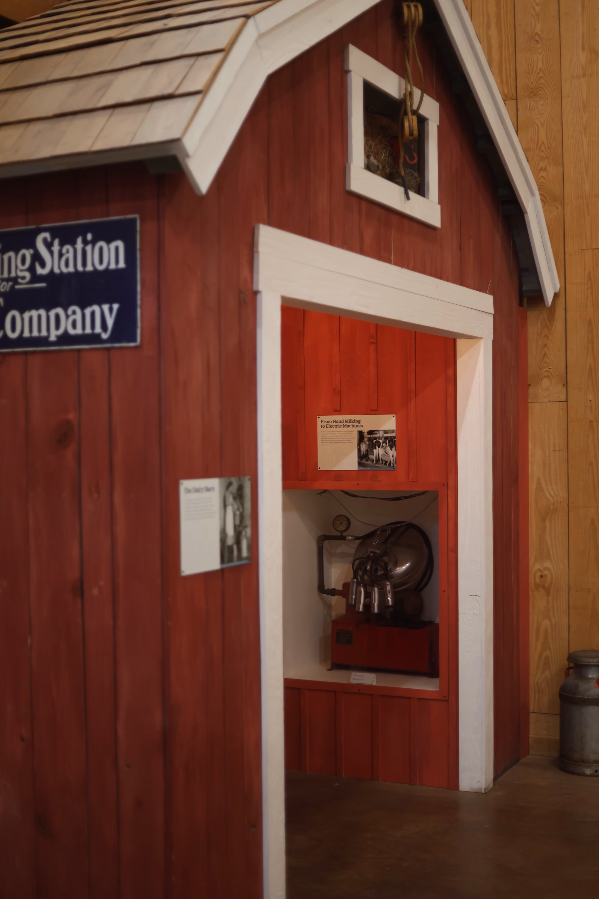 A small red wooden milking station with white trim featuring an old milking machine exhibit indoors.