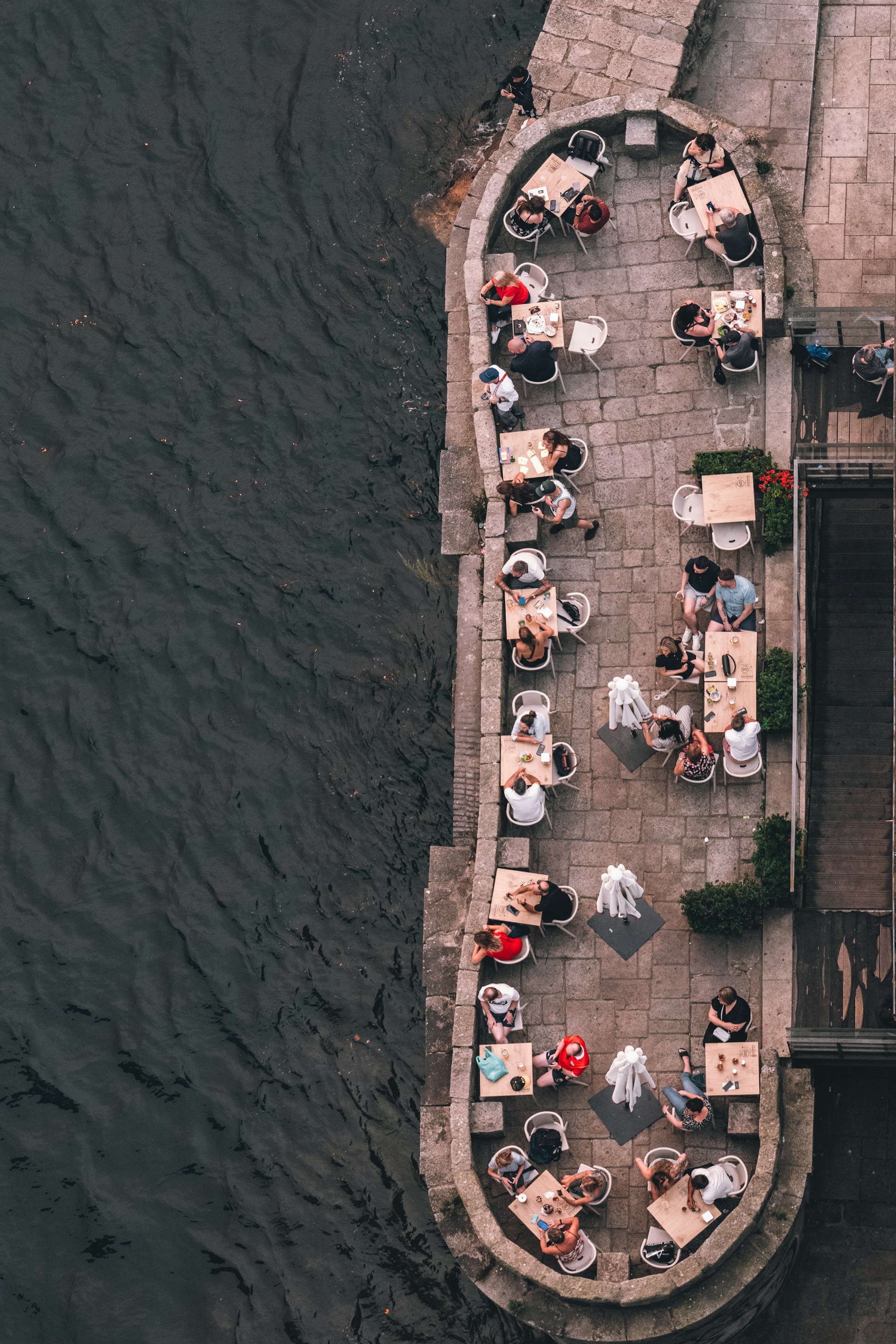 Aerial view of multiple people sitting at tables on a stone terrace next to a body of water.