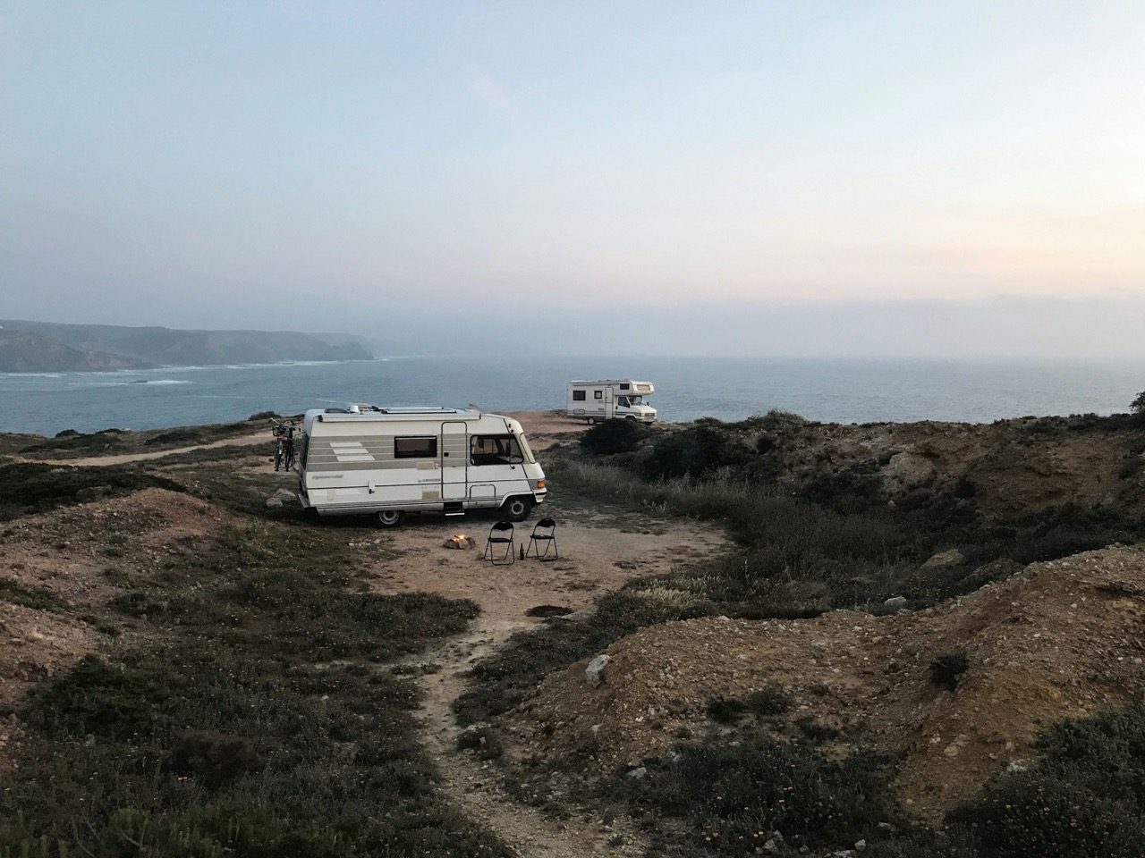 Two camper vans parked near rocky coast with two chairs and a small campfire at dusk.