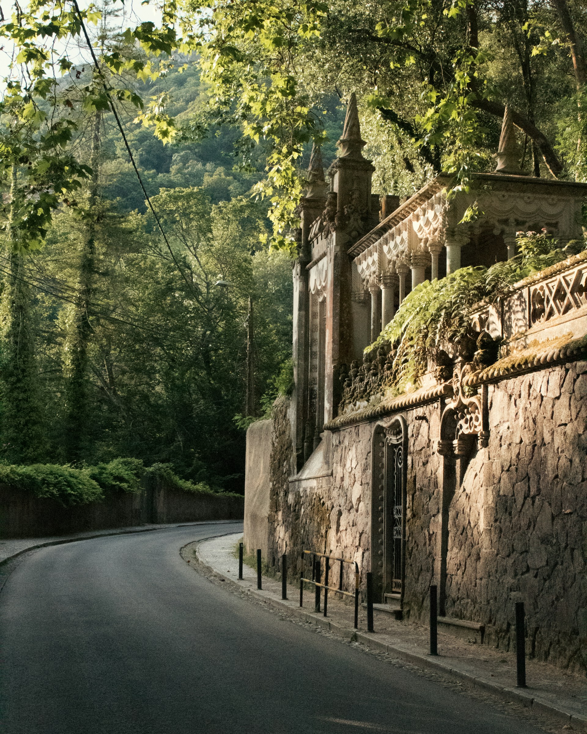 Curved empty road beside an ornate stone wall with decorative columns and lush greenery in a forested area.