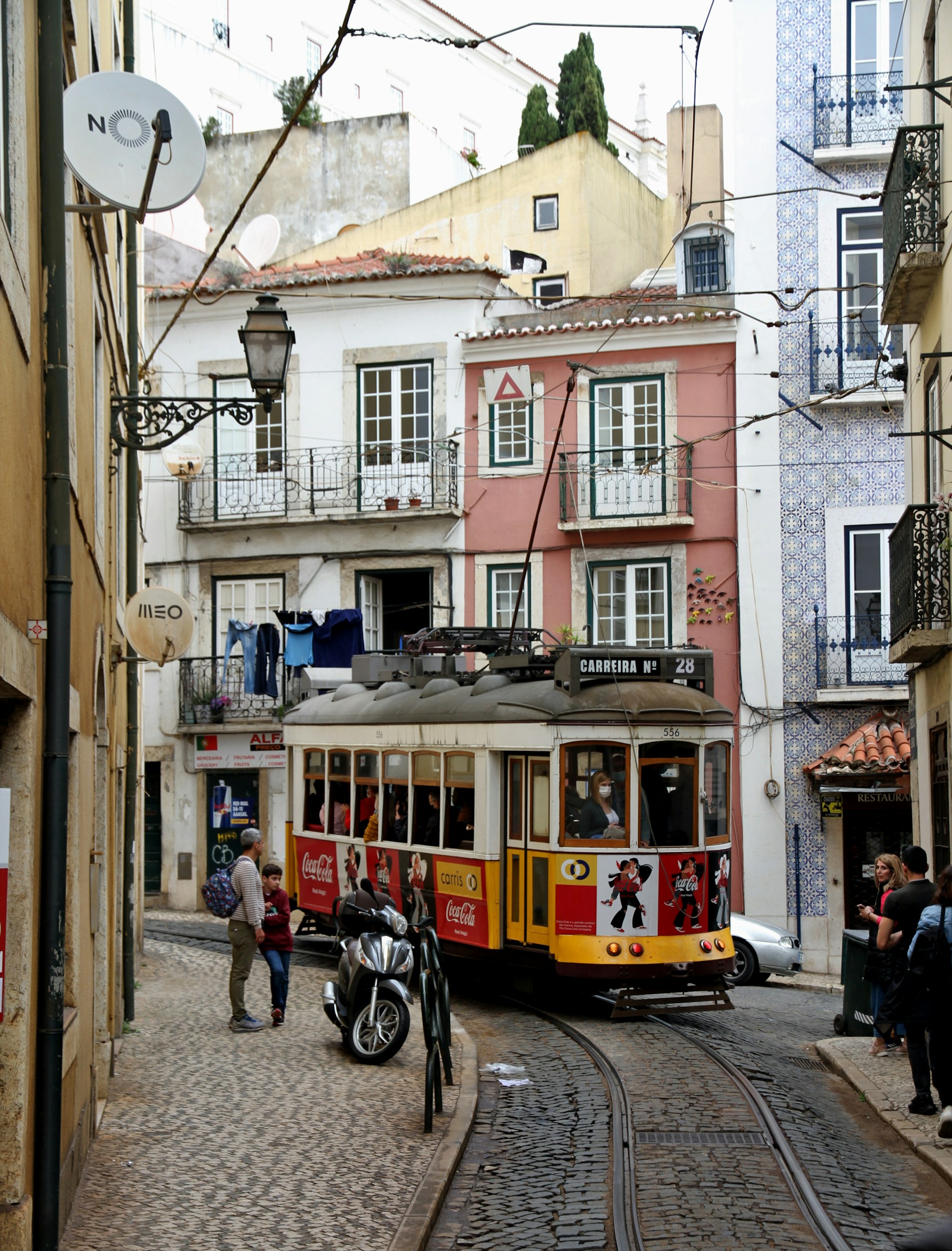 Historic yellow and red tram number 28 traveling on cobblestone street in Lisbon, surrounded by traditional buildings and pedestrians.
