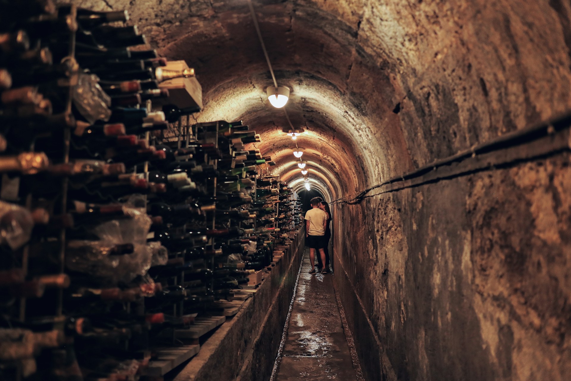 Two people standing in a dimly lit narrow wine cellar tunnel lined with racks of wine bottles on one side.
