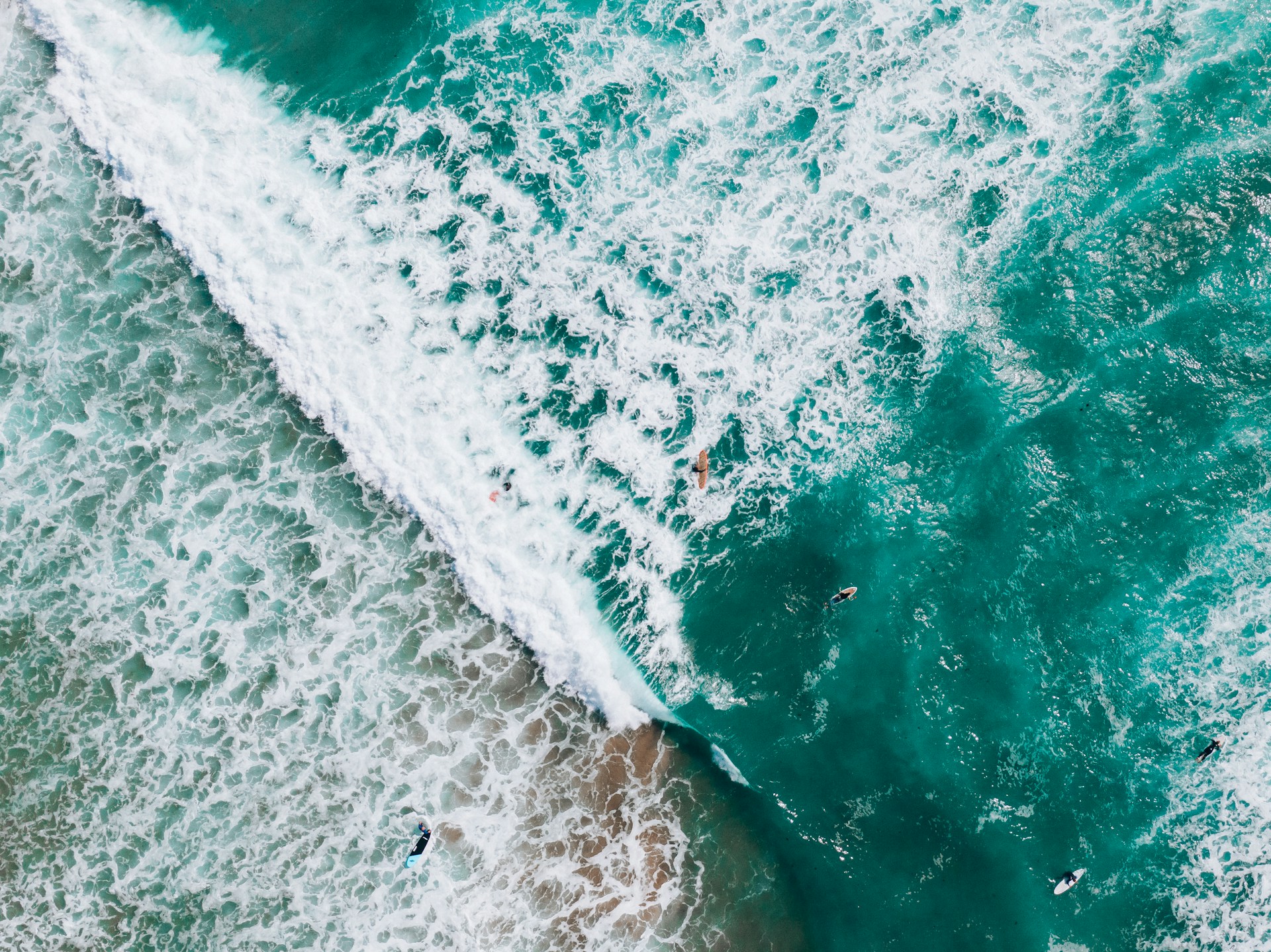 Aerial view of several surfers riding and paddling on waves in turquoise ocean water.