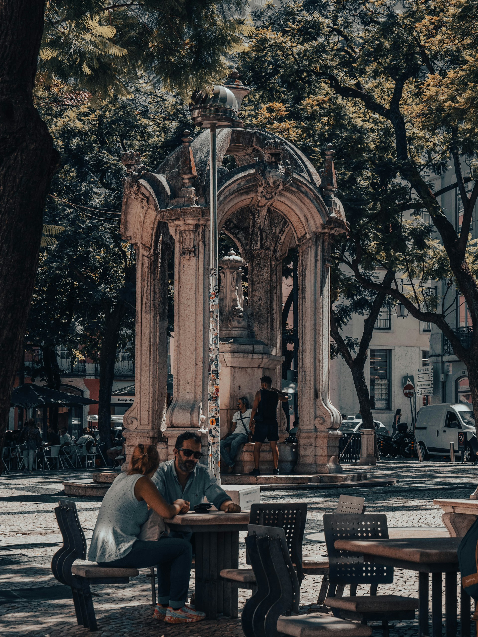 People sitting and conversing at outdoor tables near an ornate stone monument in a shaded park area.
