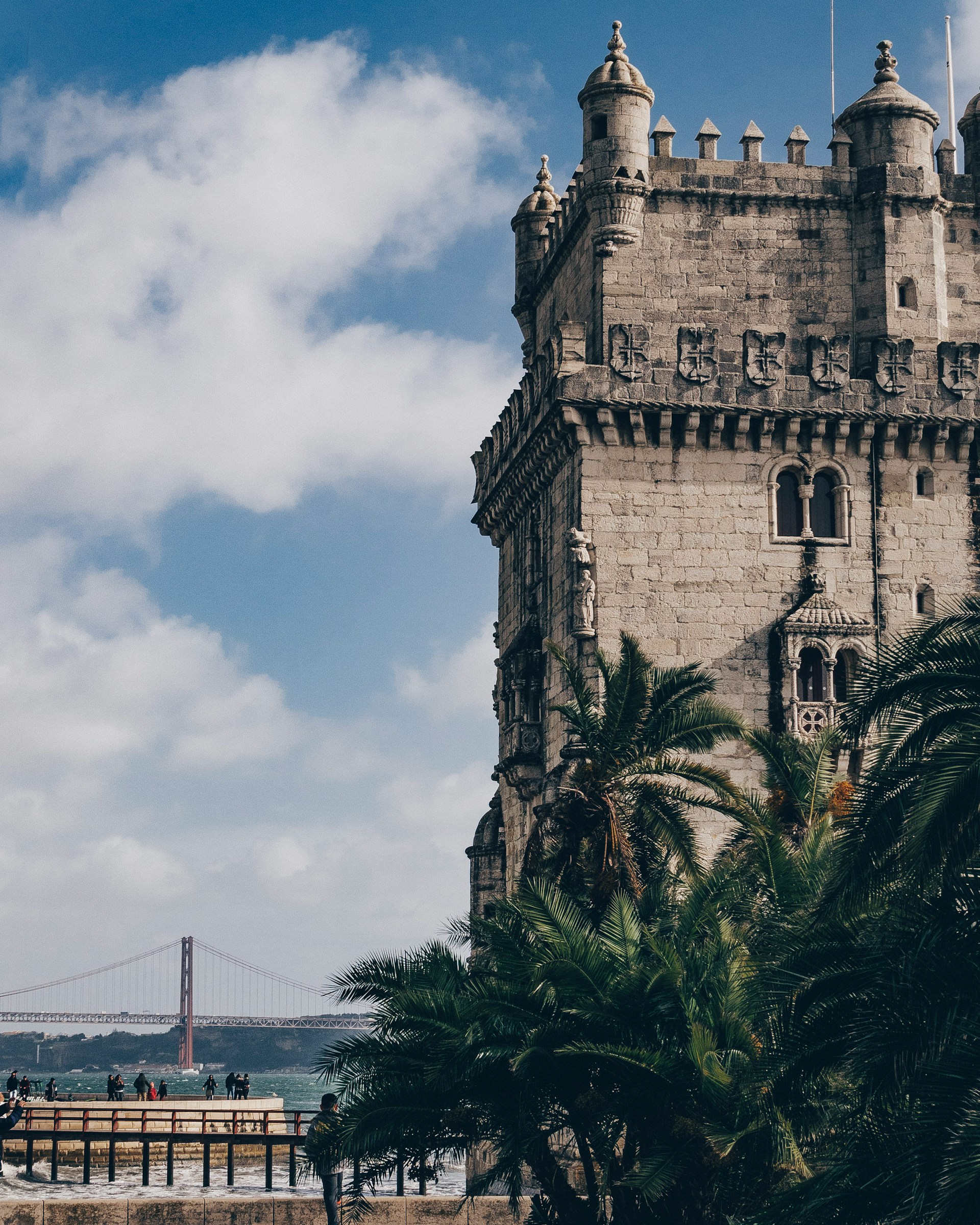 Historic stone tower with ornate details and palm trees in the foreground, with a suspension bridge and cloudy blue sky in the background.