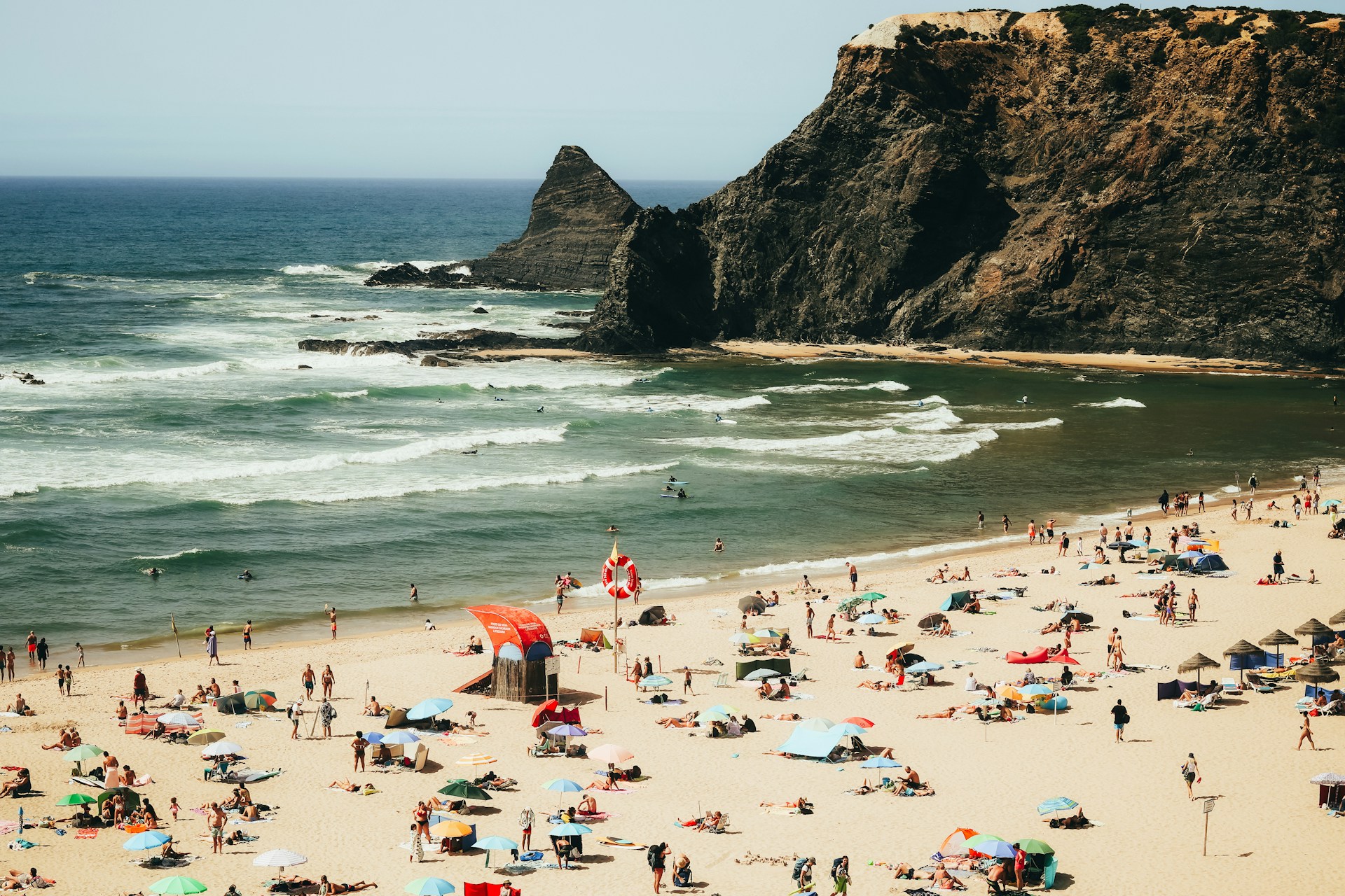 Crowded sandy beach with people sunbathing under colorful umbrellas and rocky cliffs by the ocean in the background.