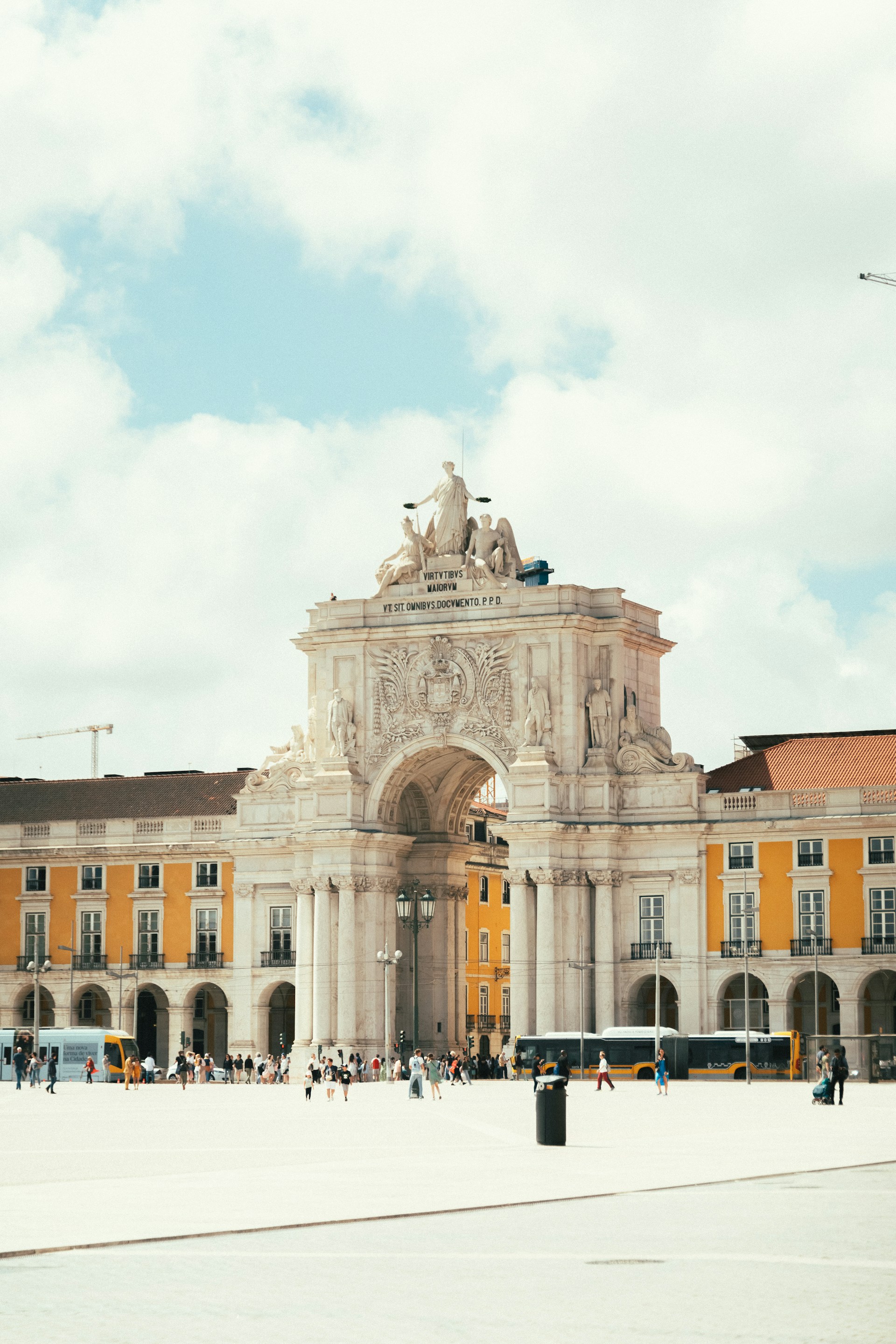 Large historic arch monument in a city square with yellow buildings and people walking around.