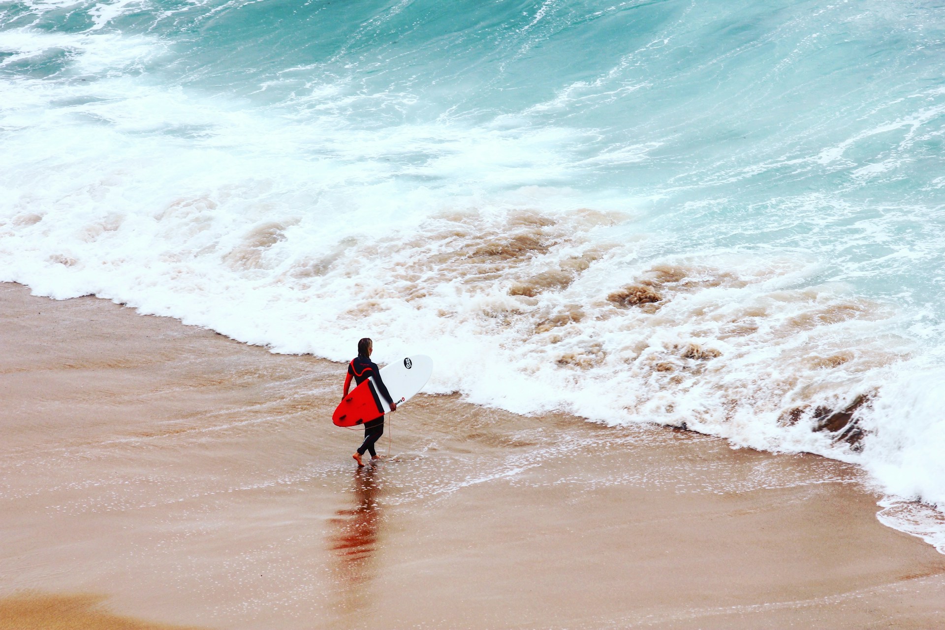 Surfer in a black wetsuit holding a red and white surfboard walking on wet sand near foamy ocean waves.